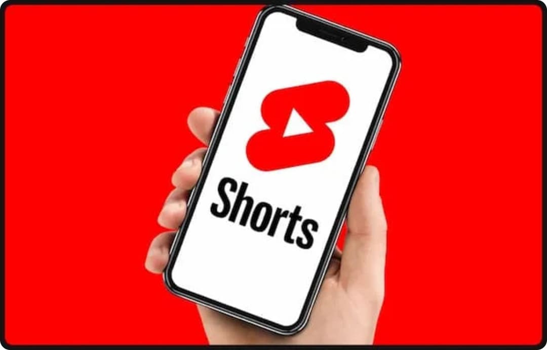 A hand holding a smartphone displaying the YouTube Shorts logo against a red background